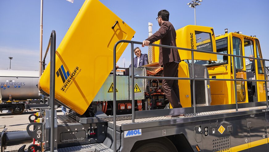 Two men are standing on a yellow two-way maneuvering device with the hood up, on the transport logistic open-air site.