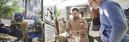 Close-up, a man stands in an exhibition hall next to a camping street sign and shows something in a catalog