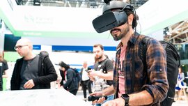 A man in a checked shirt stands at a stand with VR goggles on his head, holding two rubber handles.