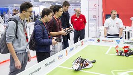 A robot arena with a few spectators and three young men with remote controls standing next to it.