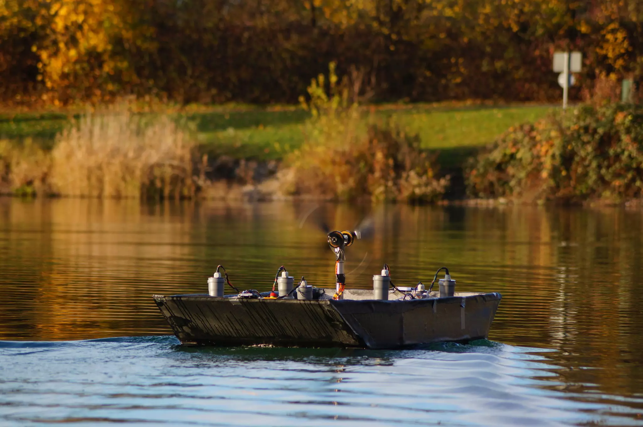 Remote-controlled boat with propeller moving on a calm lake; fall trees and greenery in the background.