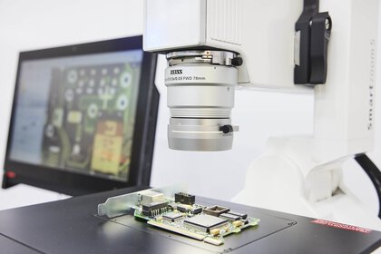 Close-up of an optical microscope with an open circuit board underneath. A monitor can be seen in the background.