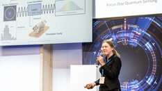Eine Frau mit Mikrofon hält einen Vortrag zum Thema Quantensensorik und steht neben einem großen Display mit Diagrammen und Text.