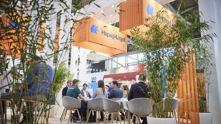 A group of people are sitting around a table, with plants to the left and right and advertising banners in the shape of orange freight containers hanging from the hall ceiling.
