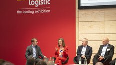 Five people are sitting on the stage at transport logistic in front of an audience. A woman in a red suit speaks straight and gestures with both hands.