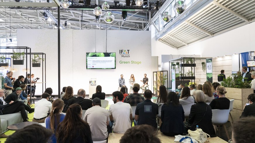 Publikum sitzt vor der IFAT Munich Green Stage, auf der drei Personen auf Hockern sitzen und sprechen.