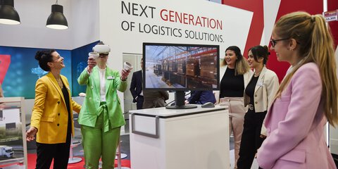 An einem Messestand der transport logistic testet eine Frau im grünen Anzug eine Anwendung mit einer VR-Brille während einige Besucher ihr dabei lächelnd zusehen.
