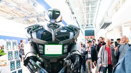 A futuristic, humanoid robot with green glowing elements stands in an exhibition hall in front of a crowd of people who are filming it.