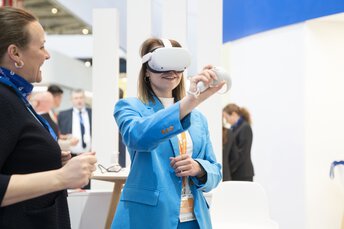 A visitor at transport logistic using a VR headset and controller while an exhibitor stands next to her.