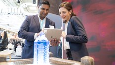 A man and a woman in business attire stand behind a stone pedestal with a kind of glowing blue crystal and look together at a tablet.