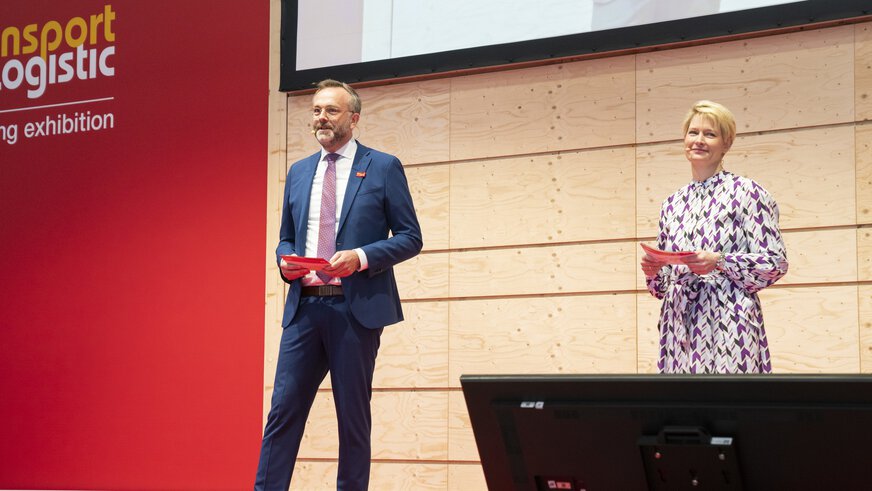 A man and a woman stand on a stage at transport logistic and speak to the audience. Behind them hangs a large screen with a live recording from the stage.