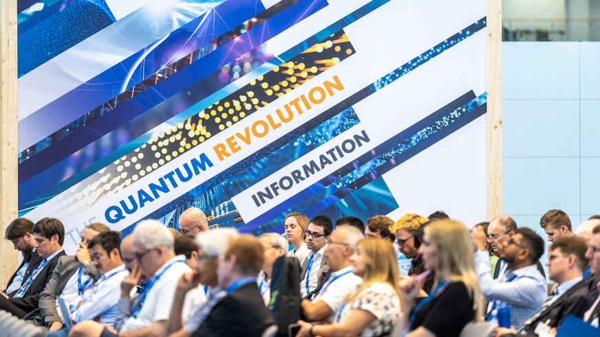Publikum auf Stuhlreihen in einer Messehalle, vor einem großen Keyvisual mit der Aufschrift 