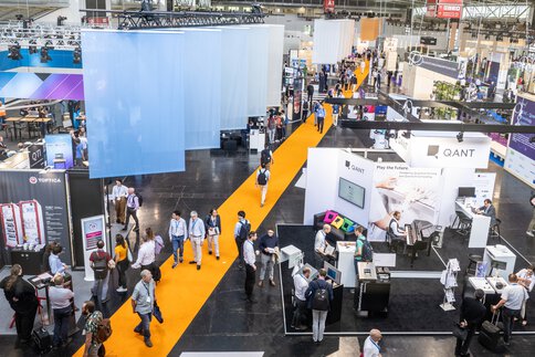 Eine geschäftige Messehalle mit Menschen, die über einen leuchtend gelben Teppich laufen, mit verschiedenen Ständen im Hintergrund.