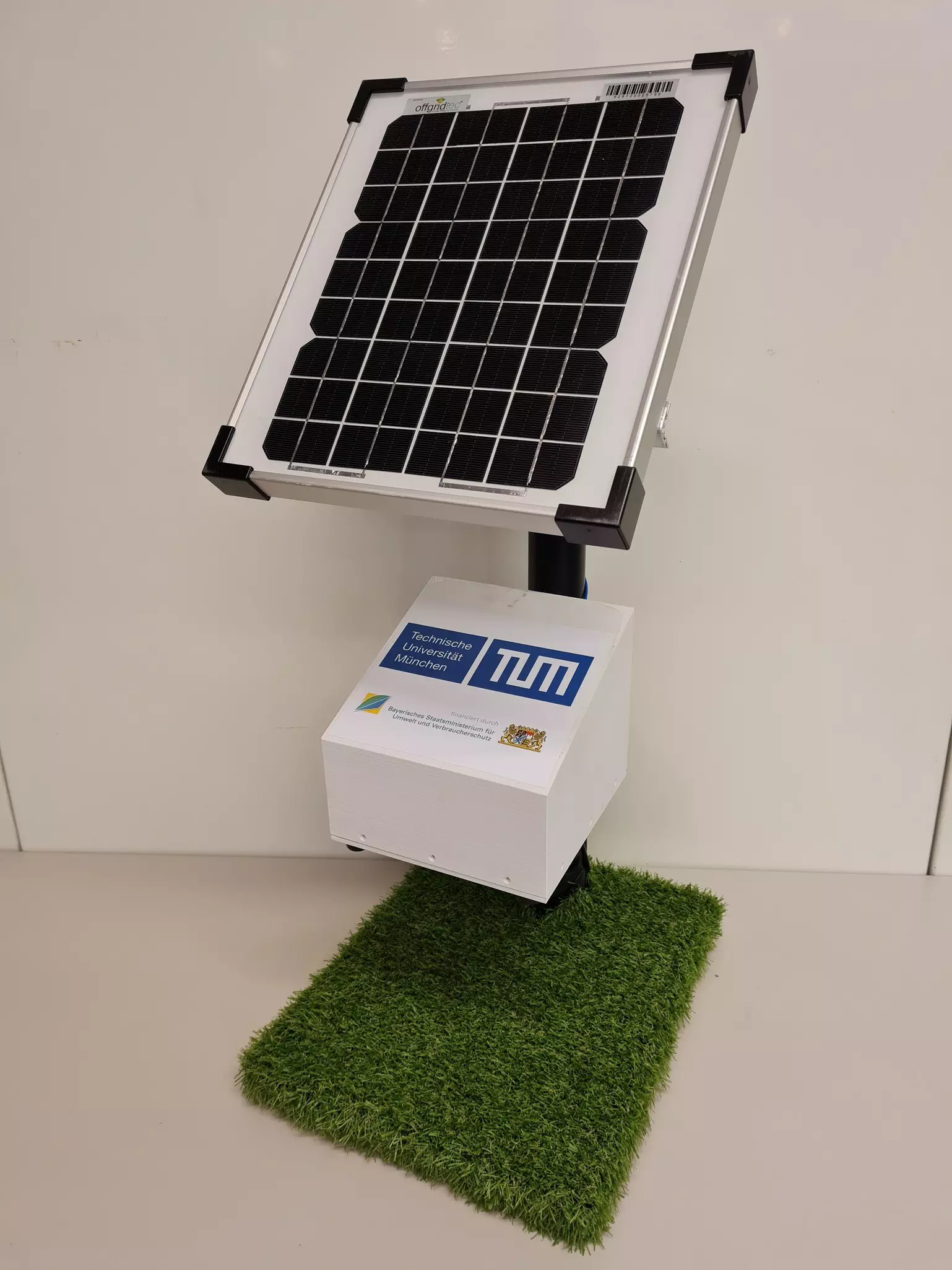 A small foam model with solar panel on artificial turf, with a white box underneath.