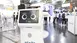 A white robot with a screen showing two eyes in an aisle between several exhibition stands.