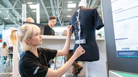 An exhibitor in a black shirt adjusting sensors on a mannequin wearing a shirt at a trade fair stand.